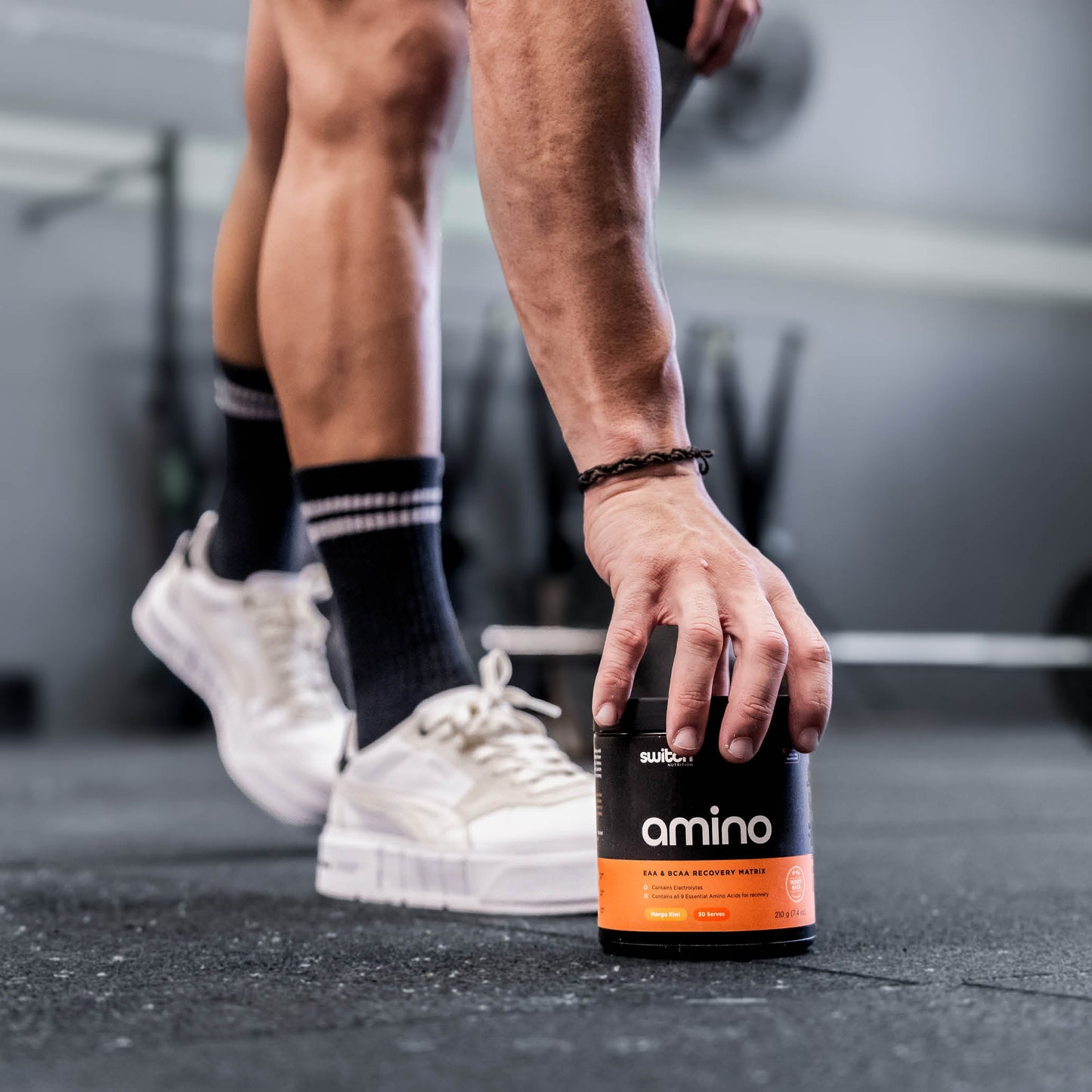 A close-up shot of a person's legs and hand in a gym setting. The person is wearing black socks and white sneakers, reaching down to pick up a container of Amino Switch EAA & BCAA Recovery Matrix supplement.