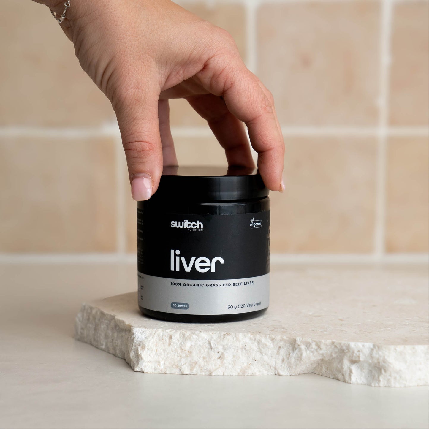 A close-up shot of a hand reaching for a container of Switch Nutrition Liver supplement, placed on a light-colored stone surface.