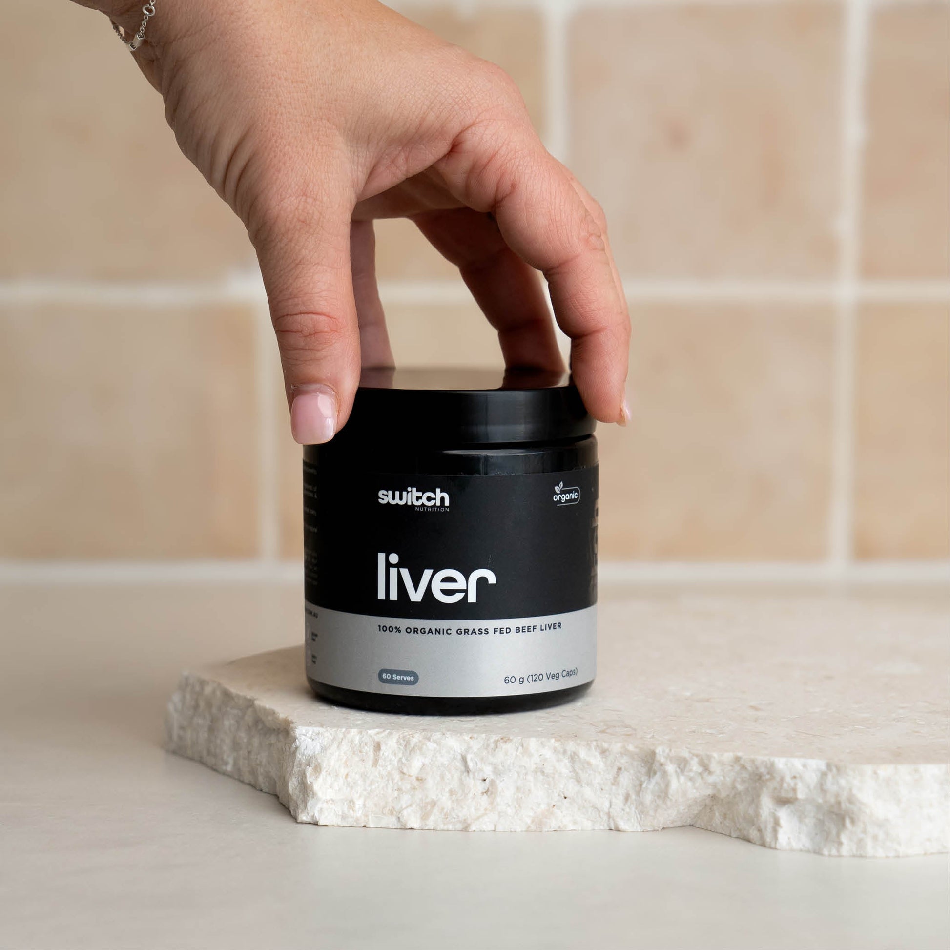 A close-up shot of a hand reaching for a container of Switch Nutrition Liver supplement, placed on a light-colored stone surface.