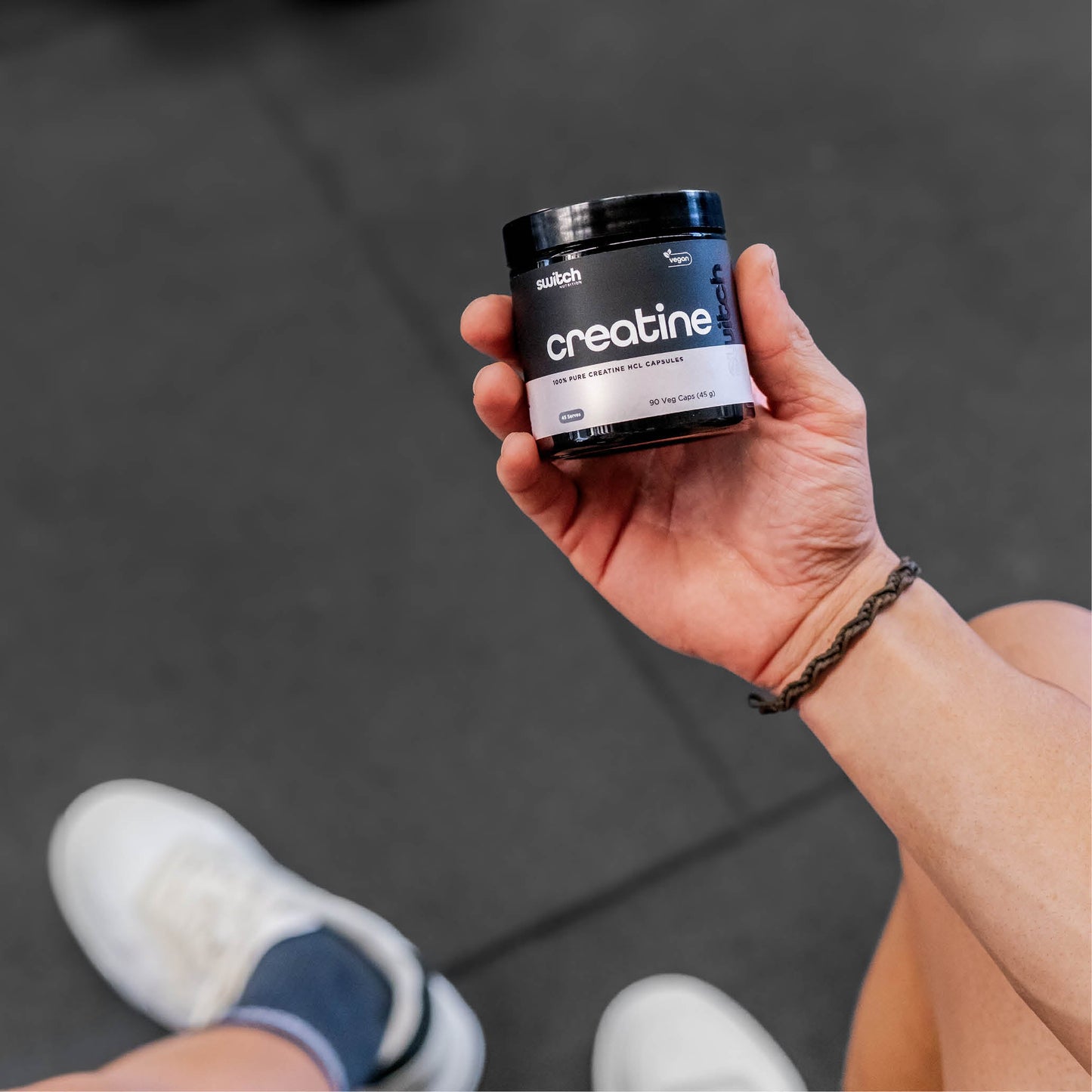A close-up shot of a person holding a container of Switch Nutrition Creatine HCL supplement, with the person's legs and white sneakers visible in the background.