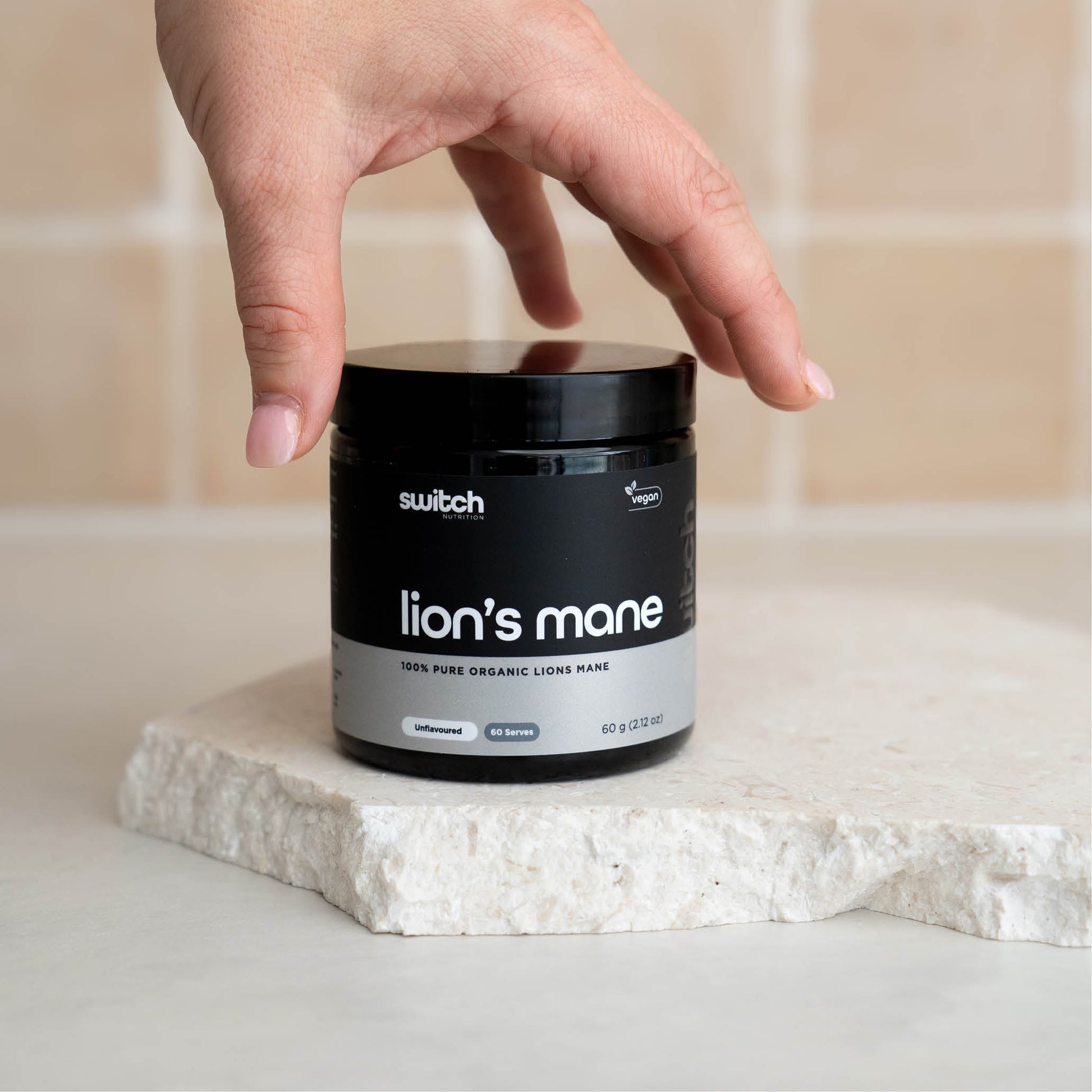 A close-up shot of a hand reaching for a container of Switch Nutrition Lion's Mane supplement, placed on a light-colored stone surface.