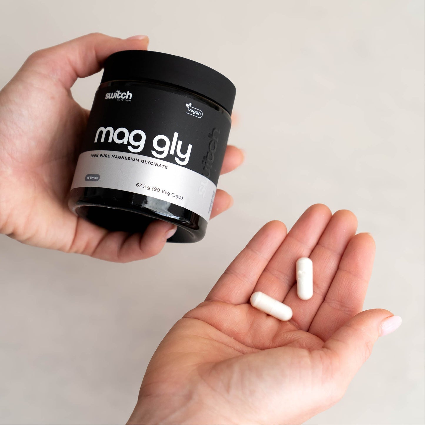 A close-up shot of hands holding a container of Switch Nutrition Mag Gly supplement, with a few capsules resting on one hand.