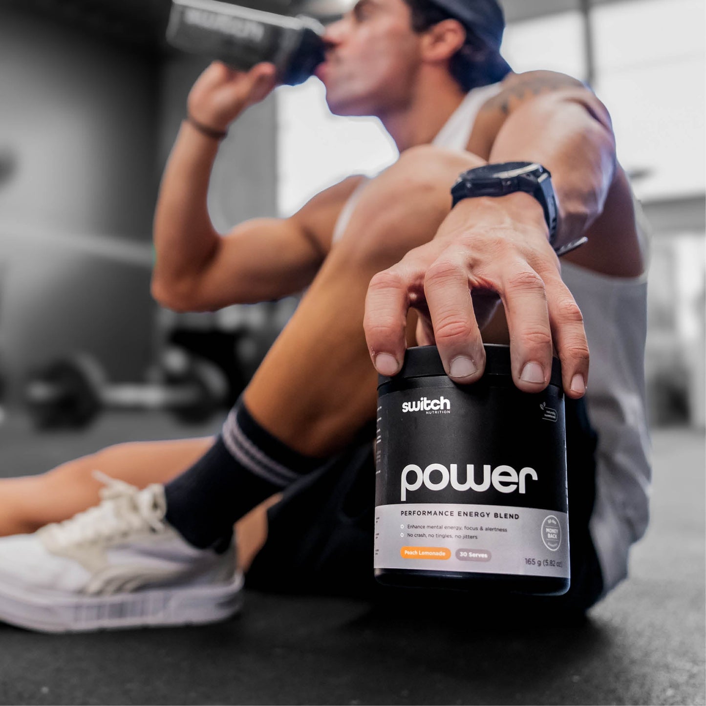 A person sitting on a gym floor, drinking from a shaker bottle and holding a container of Switch Nutrition Power Performance Energy Blend supplement, with their sneakers and gym environment visible in the background.