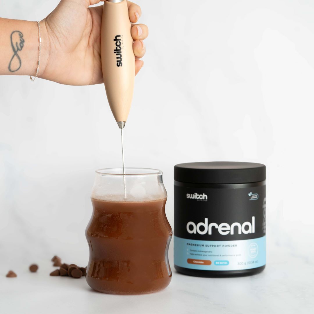Hand Blender with Adrenal Switch (Chocolate): The beige hand blender is used to mix a chocolate drink in a glass, with a tub of Adrenal Switch powder visible in the background.