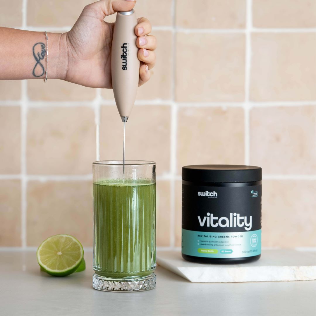 Hand Blender with Vitality Switch Greens: The beige hand blender is used to mix a green drink in a glass, with a tub of Vitality Switch greens powder visible in the background.