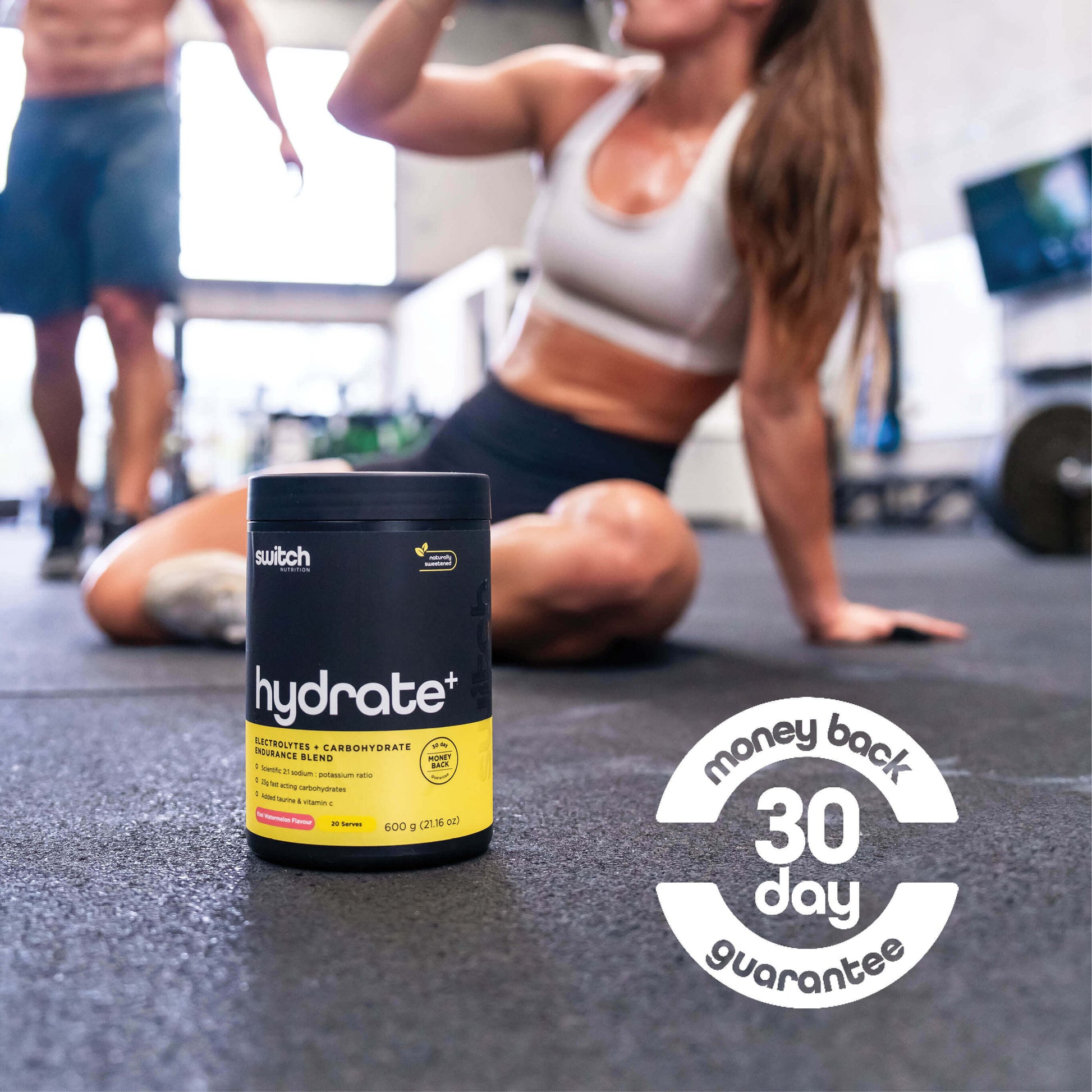 container of 'HYDRATE+' by Switch Nutrition is prominently displayed on the floor of a busy gym, with an athlete in the background in a yoga pose, which focuses on the lifestyle of active fitness enthusiasts. Emphasised at the bottom of the image is a 'money back 30-day guarantee', suggesting confidence in the product's efficacy and a commitment to customer satisfaction.