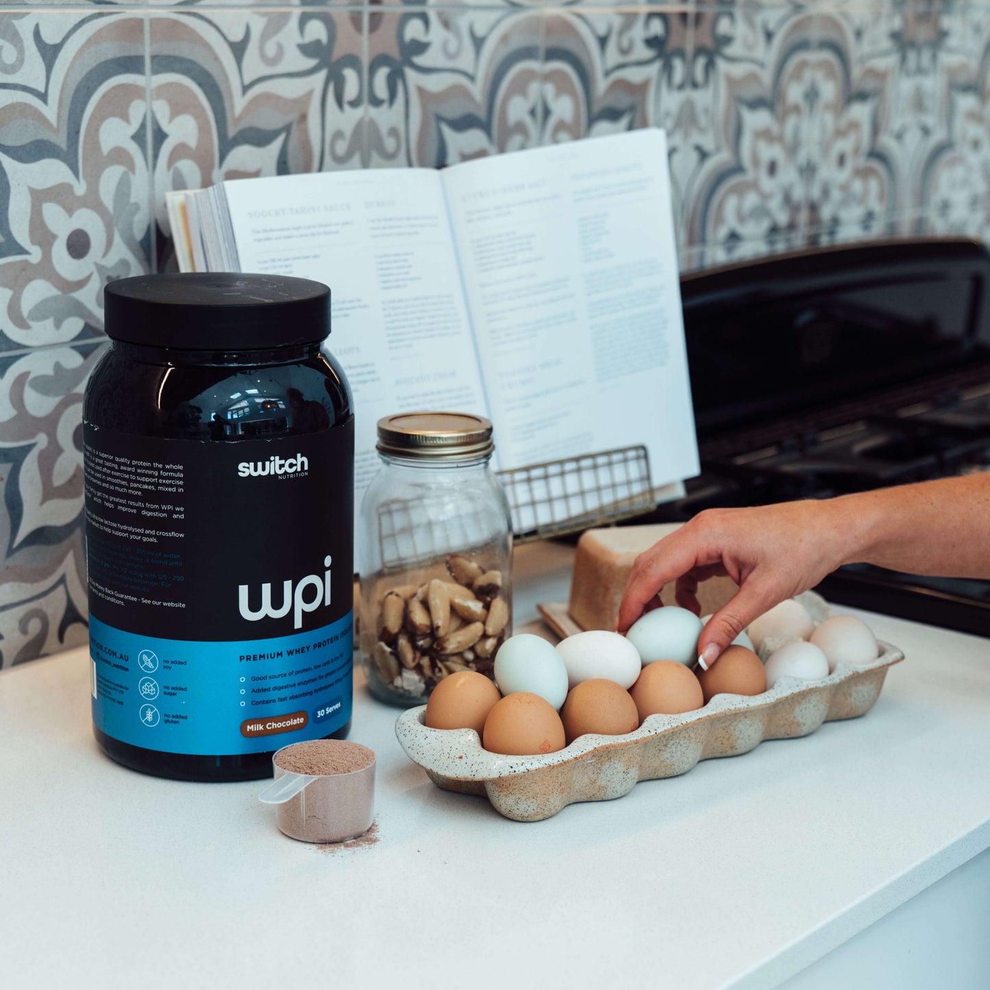 A kitchen scene with a container of WPI Switch Nutrition premium whey protein on the counter, alongside a tray of eggs, a jar of nuts, a cookbook open on a stand, and a person's hand reaching for an egg, suggesting preparation for a protein-rich meal.