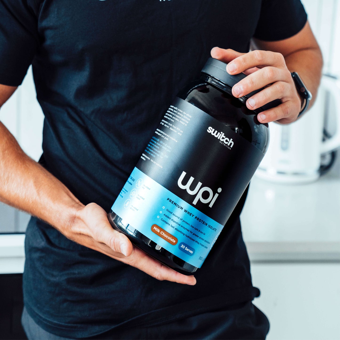 Person holding a large container of Switch Nutrition WPI, demonstrating the product's substantial size and the ease of use in a home kitchen setting.