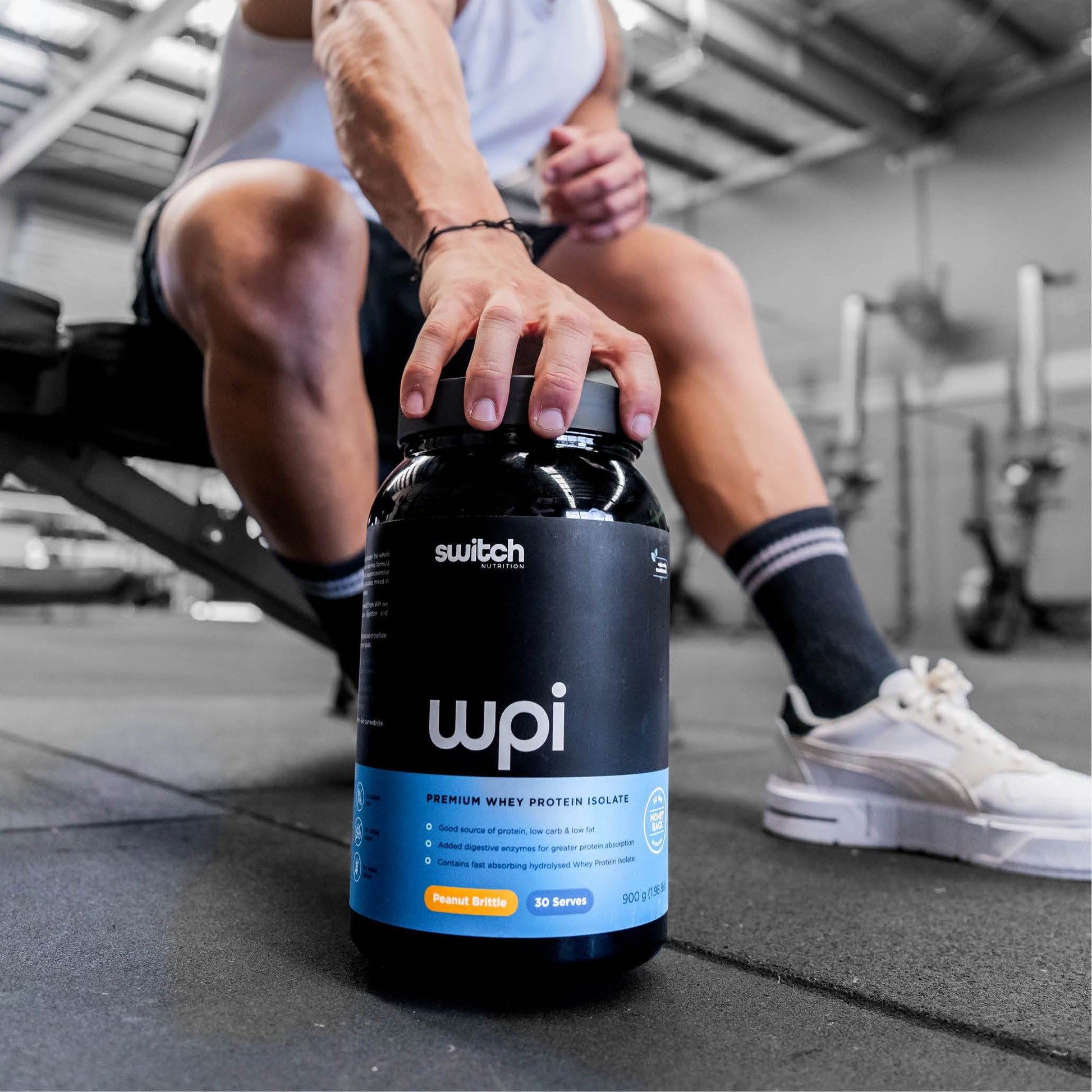 A person sitting on a gym bench, reaching for a container of Switch Nutrition WPI (Whey Protein Isolate) supplement, with their sneakers and gym environment visible in the background.