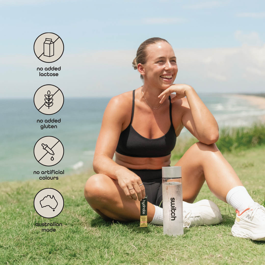 Smiling woman seated on grass by the beach with Switch Nutrition products, emphasising no lactose, gluten, artificial colours, and Australian made.