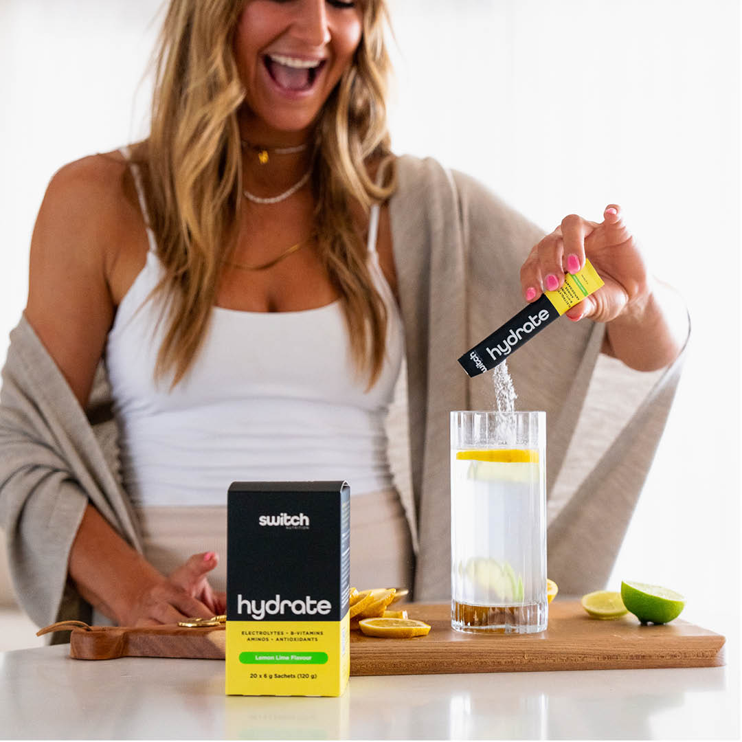 Joyful woman pouring Hydrate into a glass of water, with the product box and sachet on the kitchen counter.