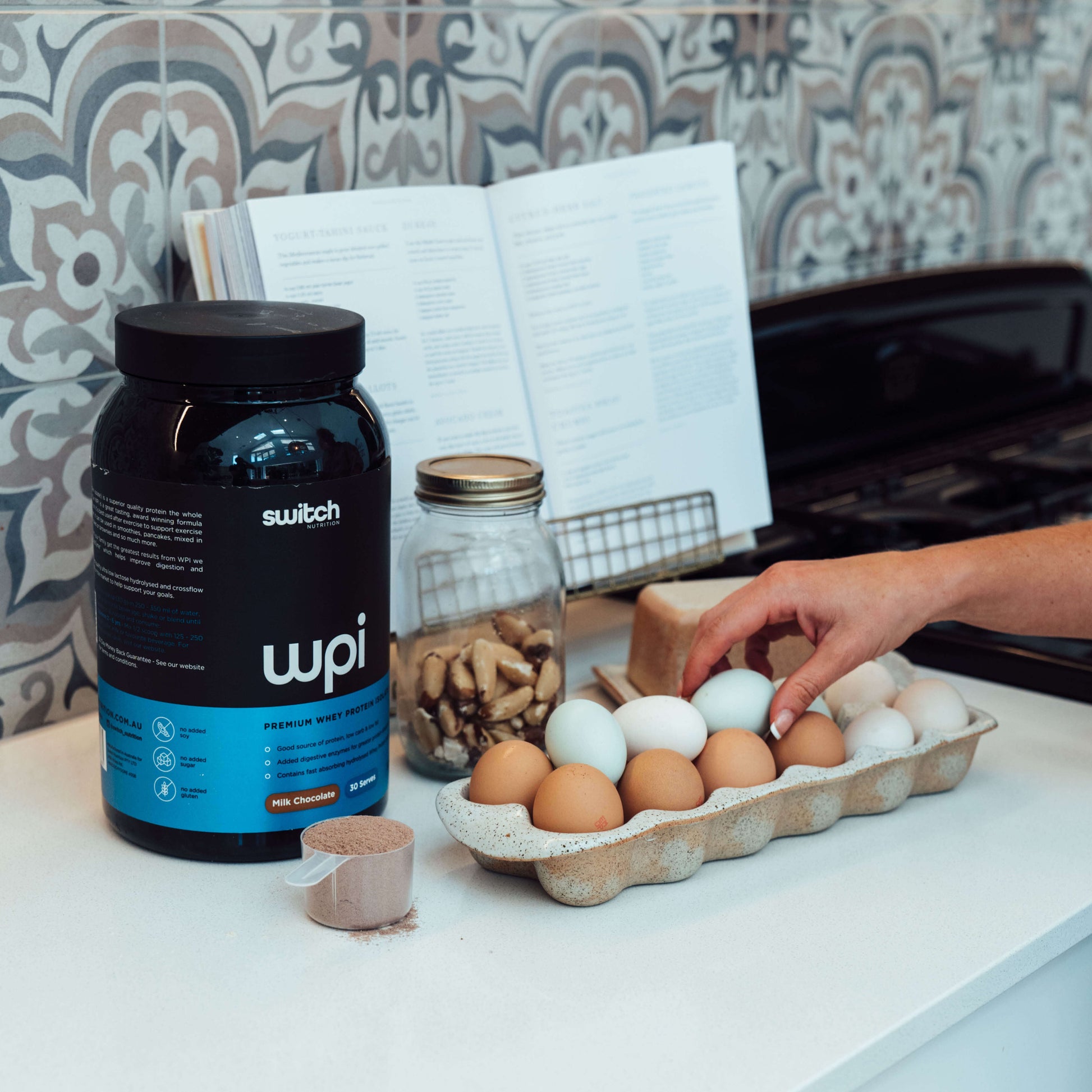 A kitchen scene with a container of WPI Switch Nutrition premium whey protein on the counter, alongside a tray of eggs, a jar of nuts, a cookbook open on a stand, and a person's hand reaching for an egg, suggesting preparation for a protein-rich meal.